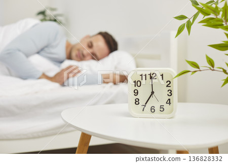 Alarm clock showing 7 AM on table with peaceful man sleeping comfortably under blanket on background Alarm clock showing 7 AM on table with peaceful man sleeping comfortably under blanket on background 136828332