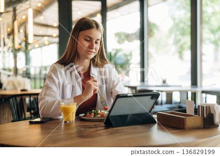 Eating, digital tablet is on the table. Young woman is in the cafe at daytime Eating, digital tablet is on the table. Young woman is in the cafe at daytime 136829199