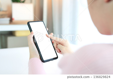 Hands of woman in pink shirt holding smart phone with white screen on white table top in a bright room background 136829824
