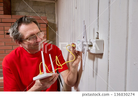 Connection of wifi router with repeater using LAN cable mature man adjusts equipment for internet distribution in rural house. Connection of wifi router with repeater using LAN cable mature man adjusts equipment for internet distribution in rural house. 136830274