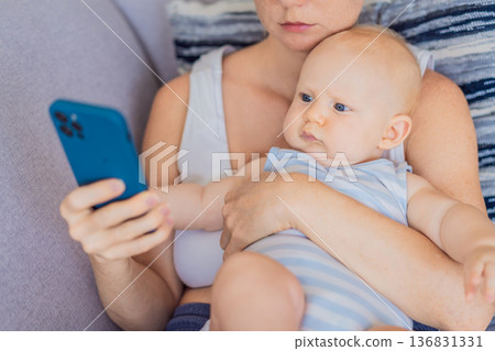 Baby sitting on mother lap while she looks at her smartphone, emotionally disconnected. Symbolic scene showing impact of screen distraction on infants and unmet emotional needs. Digital parenting and Baby sitting on mother lap while she looks at her smartphone, emotionally disconnected. Symbolic scene showing impact of screen distraction on infants and unmet emotional needs. Digital parenting and 136831331