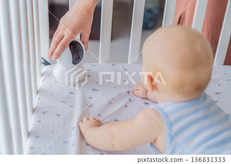 Baby monitor camera placed inside a crib where an infant is lying, ensuring safety and constant supervision. Modern parenting scene combining care and technology. Baby monitoring, security and smart Baby monitor camera placed inside a crib where an infant is lying, ensuring safety and constant supervision. Modern parenting scene combining care and technology. Baby monitoring, security and smart 136831333