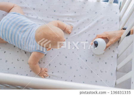 Baby monitor camera placed inside a crib where an infant is lying, ensuring safety and constant supervision. Modern parenting scene combining care and technology. Baby monitoring, security and smart Baby monitor camera placed inside a crib where an infant is lying, ensuring safety and constant supervision. Modern parenting scene combining care and technology. Baby monitoring, security and smart 136831335