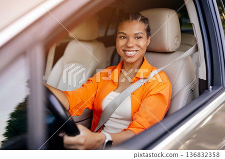 Young black woman driving car and smiling confidently at camera, wearing seatbelt Young black woman driving car and smiling confidently at camera, wearing seatbelt 136832358