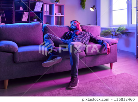 Exhausted man on couch under neon lights, evoking drugs, euphoria, and depression Exhausted man on couch under neon lights, evoking drugs, euphoria, and depression 136832460