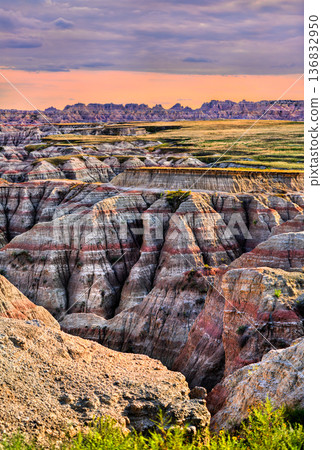Big Badlands Overlook stands in Badlands National Park, South Dakota, USA. Scenic view features colorful eroded rock formations and sedimentary layers under a cloudy sunset sky 136832950