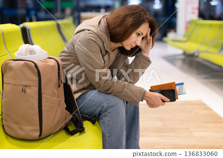 Worried woman sitting in airport terminal with backpack, holding passport and smartphone Worried woman sitting in airport terminal with backpack, holding passport and smartphone 136833060