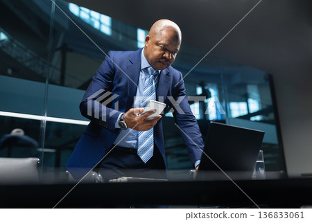 Mature black businessman standing at desk checking smartphone while working in office Mature black businessman standing at desk checking smartphone while working in office 136833061