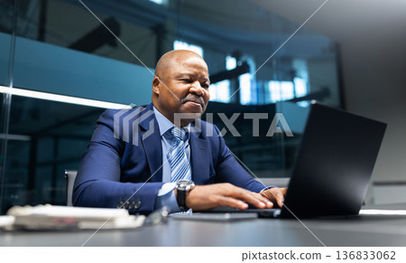 Mature black businessman smiling while working on laptop in modern office Mature black businessman smiling while working on laptop in modern office 136833062
