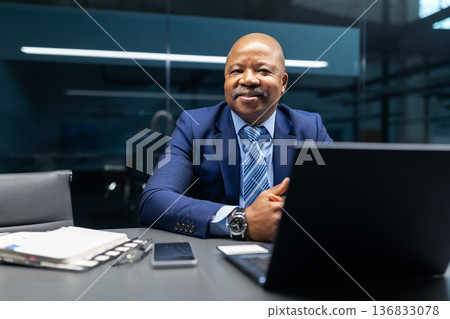 Black businessman smiling while sitting at desk with laptop in modern office 136833078