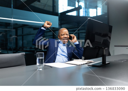 Excited black businessman celebrating success while talking on phone at office desk Excited black businessman celebrating success while talking on phone at office desk 136833090