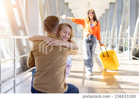 Daughter hugging father at airport while mother approaches with yellow suitcase Daughter hugging father at airport while mother approaches with yellow suitcase 136833100