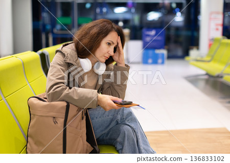 Stressed female traveler sitting in airport waiting area with headphones around neck 136833102