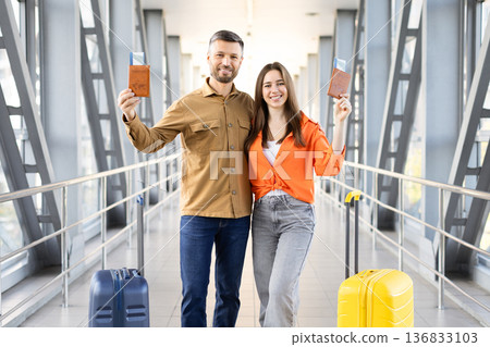 Happy couple holding passports and smiling at airport bridge, standing with suitcases 136833103