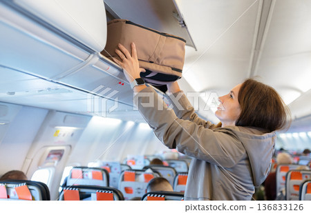 Adult caucasian woman placing backpack into airplane overhead compartment before departure Adult caucasian woman placing backpack into airplane overhead compartment before departure 136833126