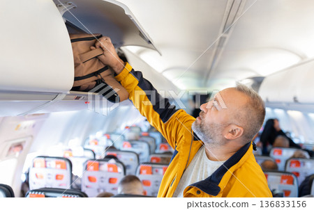 Adult caucasian male traveler placing backpack in airplane overhead compartment 136833156