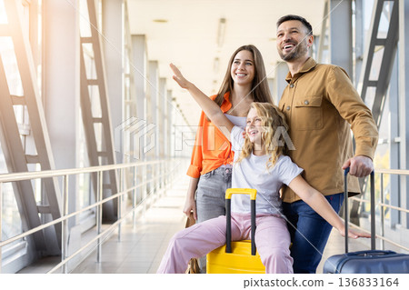 Playful girl spreading arms while sitting on suitcase at airport corridor, enjoying family holiday 136833164