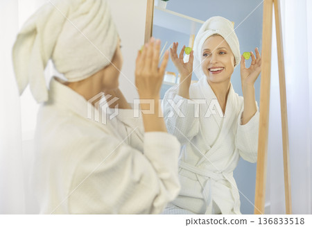 Beautiful woman wearing bathrobe and towel wrapped around her head, standing in front of mirror Beautiful woman wearing bathrobe and towel wrapped around her head, standing in front of mirror 136833518