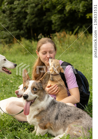 Professional dog walker young woman with pack of dogs is sitting on grass and enjoying walk animals. 136833531