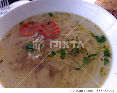 A Bowl of Soup With Clear Broth Features Noodles, Herbs, and a Slice of Tomato on Top A Bowl of Soup With Clear Broth Features Noodles, Herbs, and a Slice of Tomato on Top 136834005