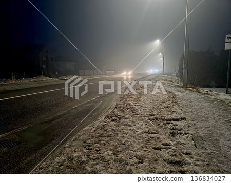 Night Road Covered in Snow With Distant Lights Shining Through Fog and Mist 136834027
