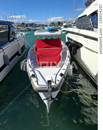A speedboat is docked in a Mediterranean marina. Bright red seating contrasts with the white boat. Luxury yachts surround the area. The scene shows clear blue sky and calm waters. 136834297