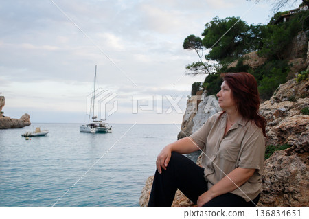 Middle aged woman sitting on the rock and enjoying sea views in Mallorca bay with yachts and boats. Calm seaside vacation in Spain. 136834651
