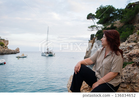 Middle aged woman sitting on the rock and enjoying sea views in Mallorca bay with yachts and boats. Calm seaside vacation in Spain. 136834652