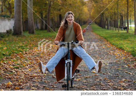Happy woman biking with legs out along autumn tree path in a forest Happy woman biking with legs out along autumn tree path in a forest 136834680