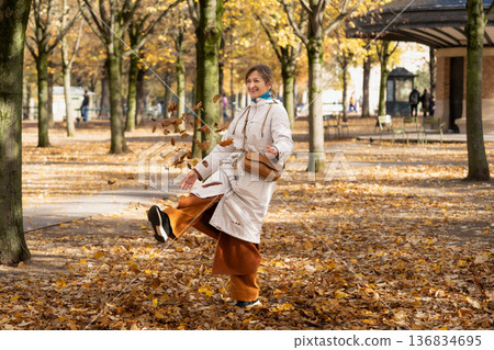 Smiling woman kicks fall leaves in sunny urban park walkway in autumn 136834695