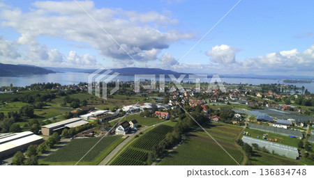 Scenic view of a lakeside village with fields and greenery during a sunny day Scenic view of a lakeside village with fields and greenery during a sunny day 136834748