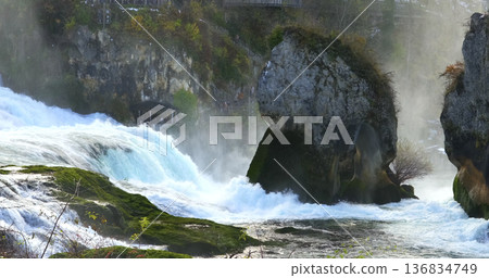 Water flows over rocks at a river during daytime in a natural setting with mist rising in the air near a forest 136834749