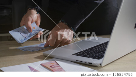 Person counts cash while using a laptop on a wooden table in an indoor setting during daytime Person counts cash while using a laptop on a wooden table in an indoor setting during daytime 136834795