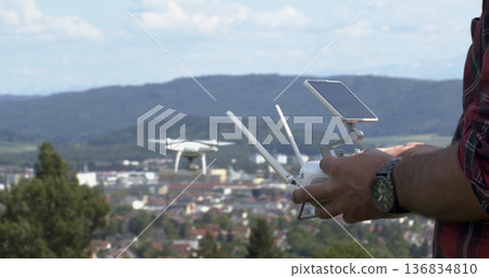 Person operates a drone above a city in the daytime with hills in the background Person operates a drone above a city in the daytime with hills in the background 136834810