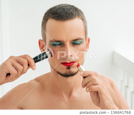 gay man with makeup and cosmetics on face. A transgender male with eye shadows paints lips with red lipstick and applies a blush brush to his face on a white background gay man with makeup and cosmetics on face. A transgender male with eye shadows paints lips with red lipstick and applies a blush brush to his face on a white background 136841768