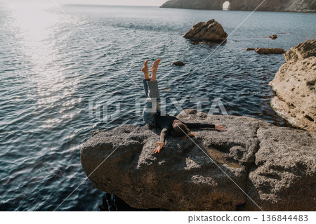 Woman sea rock. Young woman lies on a large coastal rock with her legs raised during a peaceful morning by the ocean Woman sea rock. Young woman lies on a large coastal rock with her legs raised during a peaceful morning by the ocean 136844483