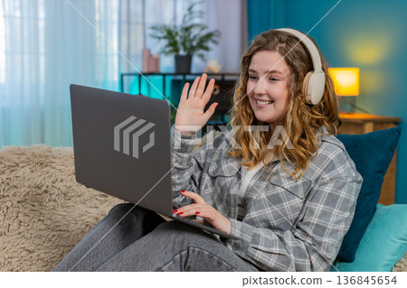Woman sits on sofa wearing headphones focused looking at laptop during video call or online course 136845654