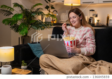 Adult woman with laptop and popcorn watches trailer movie online laughs comments comedy fun at night Adult woman with laptop and popcorn watches trailer movie online laughs comments comedy fun at night 136845659