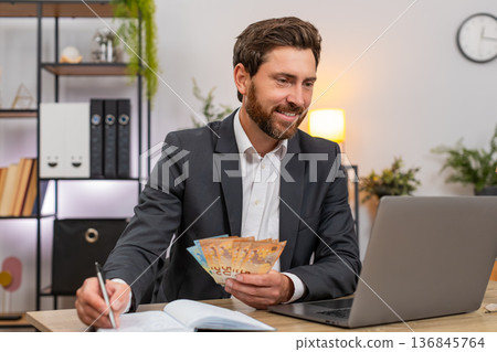 Middle-aged businessman counts cash euros enters budget on laptop writes notes in notebook and plans Middle-aged businessman counts cash euros enters budget on laptop writes notes in notebook and plans 136845764