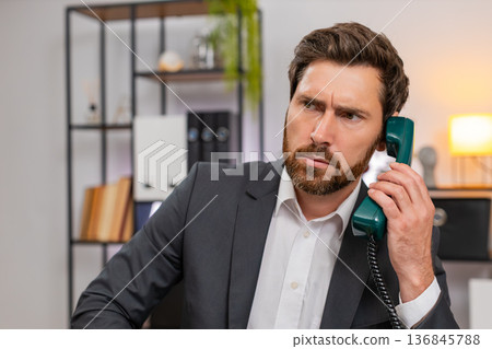 Middle-aged businessman lifts retro wired telephone and makes client call listening closely calmly Middle-aged businessman lifts retro wired telephone and makes client call listening closely calmly 136845788
