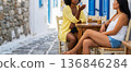 conversation between two women at a table over their favorite iced coffee drink at a street cafe on an old street of a city on the Greek or Italian islands in the Mediterranean, copy space for text 136846284