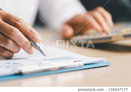 Hand writing notes in a notebook beside a calculator on office desk for budgeting and accounting. Personal finance planning, expense tracking, tax calculation, soft focus background, copy space. 136847177
