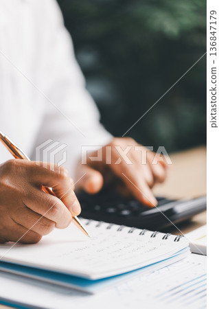 Hand writing notes in a notebook beside a calculator on office desk for budgeting and accounting. Personal finance planning, expense tracking, tax calculation, soft focus background, copy space. 136847179