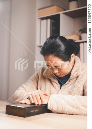 Asian woman praying with closed eyes and hands together above an open book, warm indoor light and soft focus, expressing faith, devotion, mindfulness and peaceful reflection at home. Asian woman praying with closed eyes and hands together above an open book, warm indoor light and soft focus, expressing faith, devotion, mindfulness and peaceful reflection at home. 136847200