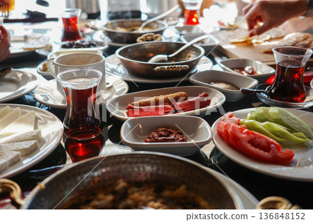 Rich traditional Turkish breakfast spread with cheeses, olives, sausages, tomatoes, cucumbers, pastries, jams and black tea in tulip glasses, hands serving food 136848152