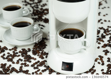 White drip coffee maker with a full cup of fresh coffee and scattered beans. White drip coffee maker with a full cup of fresh coffee and scattered beans. 136848574