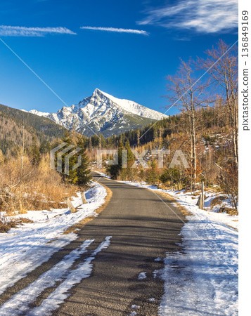 Krivan mountain in High Tatras National Park, Slovakia, Europe. A winding mountain road with snow-covered sides leads towards a majestic, snow-capped peak under a bright blue sky on a sunny day. 136849169