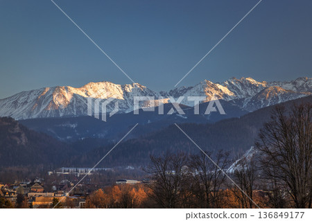 Snow-capped mountains rise majestically over a city nestled among rolling hills and forests under a clear sky. Zakopane town at the foot of the Tatra Mountains in Poland, Europe. Snow-capped mountains rise majestically over a city nestled among rolling hills and forests under a clear sky. Zakopane town at the foot of the Tatra Mountains in Poland, Europe. 136849177