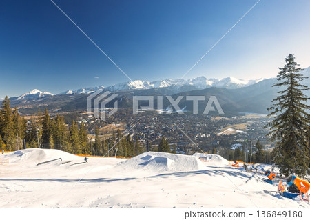 A panoramic view shows a snowy mountain range under a clear blue sky, with a ski slope in the foreground and city afar. Zakopane town at the foot of the Tatra Mountains in Poland, Europe. A panoramic view shows a snowy mountain range under a clear blue sky, with a ski slope in the foreground and city afar. Zakopane town at the foot of the Tatra Mountains in Poland, Europe. 136849180