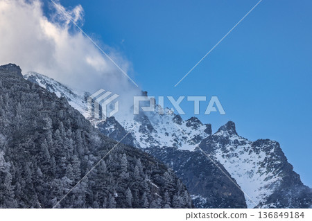 Scenic landscape featuring a snow-covered mountain partially obscured by clouds, with a dense forest covering the lower slopes, under a clear blue sky. High Tatras National Park, Slovakia, Europe. 136849184